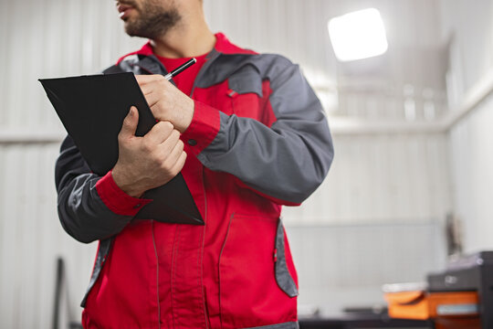 Mechanical technician taking notes on a clipboard, attired in a distinctive red and gray uniform, set against the backdrop of a functional auto maintenance garage for professional service
