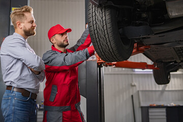 A car mechanic and a customer are inspecting a vehicle in a professional auto shop. The mechanic...