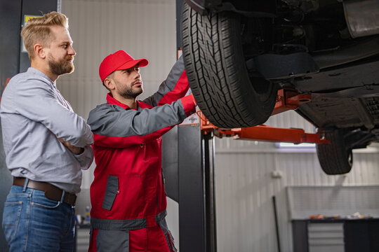 A car mechanic and a customer are inspecting a vehicle in a professional auto shop. The mechanic explains repair details while showing car parts to the attentive customer.