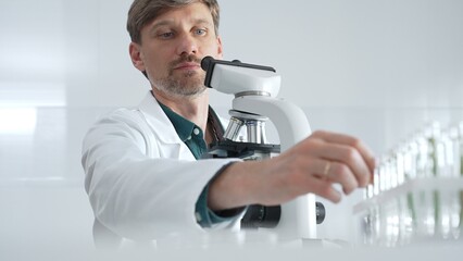 Adult male scientist wearing white mask, lab coat and protective glasses using microscope, doing...