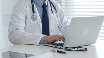 Male doctor in lab coat and stethoscope sitting at desk typing on laptop, holding glasses and pen...