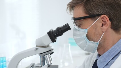 Scientist wearing mask and gloves using microscope doing scientific research in a laboratory with...