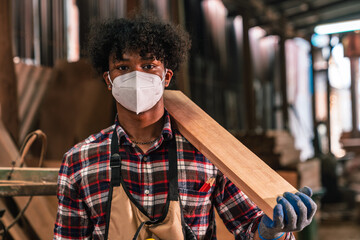 Portrait Of Young African American Carpenter Holding Wood Plank In Timber Workshop, Wearing Apron...