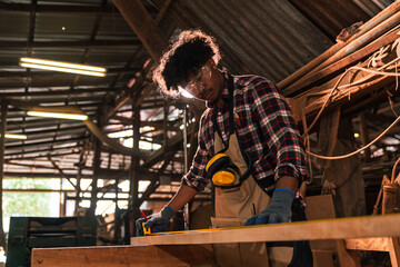 Young African American Carpenter Measuring Wood Plank with Tape and Pencil, Working in Sawmill...
