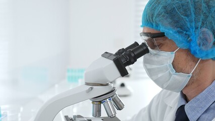 Senior male scientist wearing protective mask, cap and gloves using microscope in laboratory,...