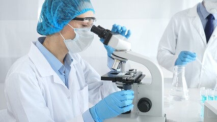 Fototapeta premium Woman scientist wearing blue protective gear is examining specimen under microscope while laboratory assistant handles glassware in medical investigation at the background. Medicine, and science