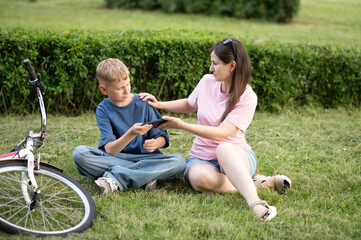 Mother and son looking at smartphone while sitting on grass next to bicycle. Teenagers children...