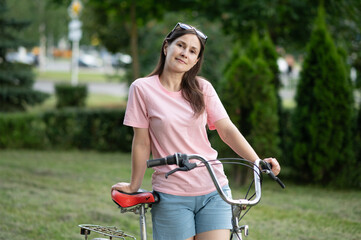 Woman standing with bicycle, holding handlebars and looking confident. Casual summer clothes, green...