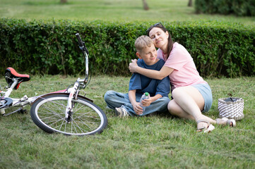 Mother lovingly hugging her son as he sits on the grass with a tablet and bicycle nearby. Tender...