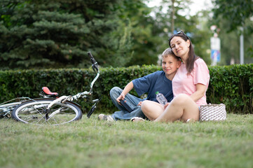 Mother and son hugging on grass near a bicycle. Loving family moment, both smiling and relaxed, in...