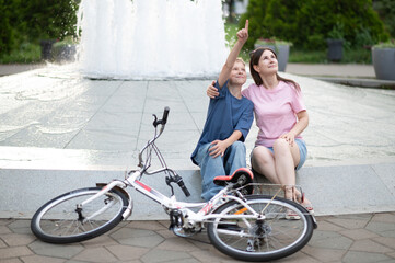 Mother and son sitting near a fountain with a bike, pointing up and smiling. Casual summer clothes,...