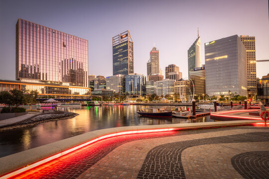 View of modern buildings mirrored in the tranquil waters, red lights lining the walkway, creating a vibrant contrast against the twilight sky, Perth, Western Australia, Australia.
