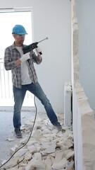Man construction worker demolishing a wall with a hammer drill in protective gear, creating construction debris on the surrounding floor surface. Renovation concept