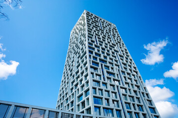 Modern building with geometric facade against blue sky, clouds. Sunny day emphasizes height, style, windows, lines, structure. Tall skyscraper rushes into clear sky