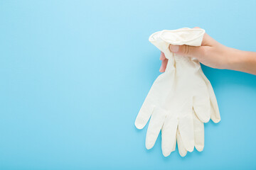 Young adult woman hand holding and showing white latex protective gloves on light blue wall background. Pastel color. Closeup. Empty place for text. Side view.