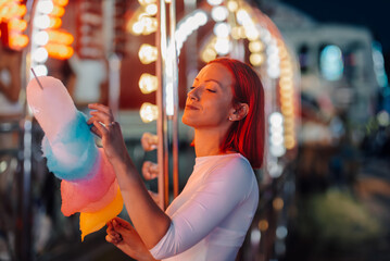 Redhead woman enjoying cotton candy at amusement park night lights