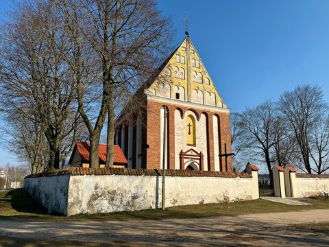  St. Anne Church in Skaruliai, Jonava, Lithuania. Red-brick church with tall arched windows and gable roof. Surrounded by leafless trees within a white stone wall. 