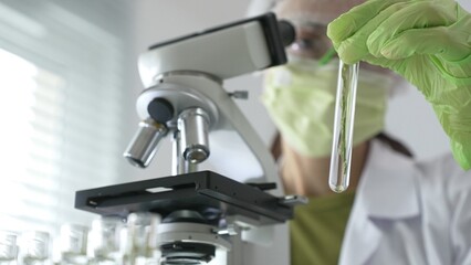 Female scientist in green protective mask and gloves holding test tube with plant sample, researching botany and medical applications using a microscope. Medicine, health care and science concept