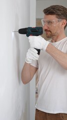 Middle aged male worker wearing white t-shirt and protective gloves, is drilling hole in a wall...