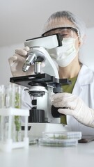 Female scientist wearing white protective gear using microscope, analyzing test tubes in...
