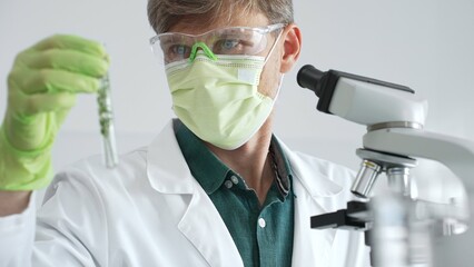 Adult male scientist wearing green mask, gloves and goggles using microscope and analyzing sample...