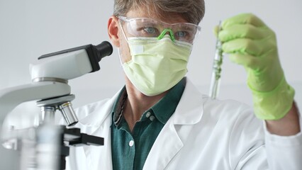 Adult male scientist wearing green mask, gloves and goggles using microscope and analyzing sample...