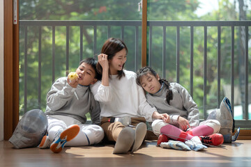 The fencers' families rest on the floor as they wait for the next round of the competition. Mother, son and daughter sit against a glass wall under the warm sunlight.