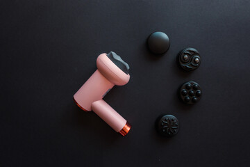 Pink massage gun with soft head and four different black attachments arranged on dark background....