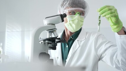 Adult male scientist wearing green protective gear analyzes plant samples in test tubes and uses a microscope in a modern laboratory for botanical research. Medicine, healthcare and science concept - Powered by Adobe