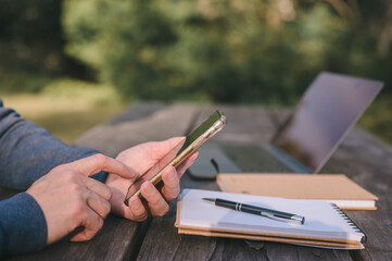 Close-up of a man's hands using a smartphone while sitting at a wooden outdoor table in a park. A...