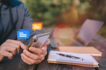 Close-up of a man's hands using a smartphone while sitting at a wooden outdoor table in a park. A...