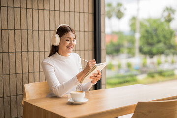 Young woman wearing headphones taking notes and drinking coffee in cafe