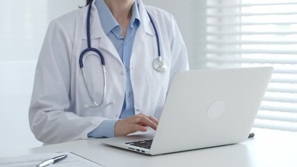 Female doctor physician working at desk, typing on laptop, medical records and pen nearby, bright window with blinds illuminating clinical workspace. Medicine and health care concept - Powered by Adobe