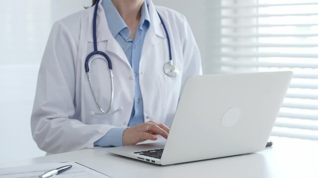 Female doctor physician working at desk, typing on laptop, medical records and pen nearby, bright window with blinds illuminating clinical workspace. Medicine and health care concept - Powered by Adobe