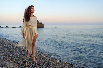 Barefoot woman strolling sandy shoreline, sunset casting warm glow, experiencing peaceful moment...