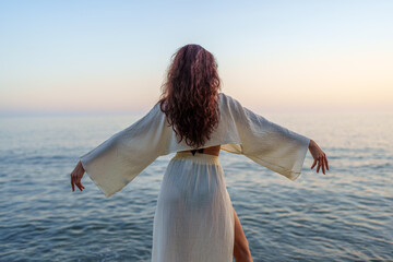 Young woman spreading arms, facing ocean, long brown hair flowing, white outfit, sunset landscape,...