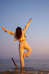 Professional woman balancing on leg near ocean during sunset, working laptop beside her, enjoying...