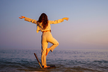 Professional woman balancing on chair, performing yoga pose during golden sunset over calm sea,...
