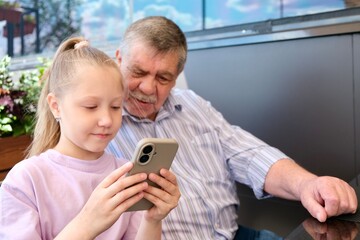 Granddaughter showing smartphone to grandfather. Portrait of a cute girl playing or typing on her smartphone while sitting in a cafe with her grandfather.