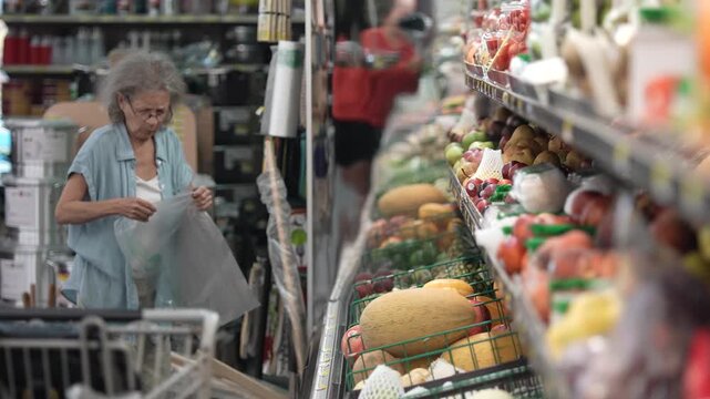 An elderly senior woman is selecting fresh produce while grocery shopping in a local store. She appears focused as she navigates through the aisles.