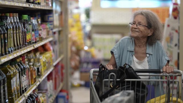 An elderly woman pushes a shopping cart as she thoughtfully looks at product shelves in a grocery store, contemplating her meal options while shopping.