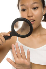 Asian woman surprised while inspecting her hand with magnifying glass, studio shot on white...