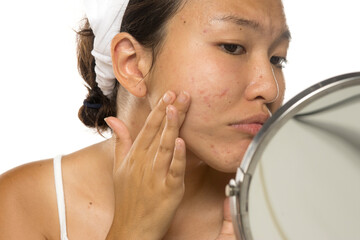 Asian woman touching her cheek while examining skin in mirror, studio shot on white background.