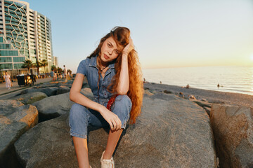Lifestyle woman in modern boho-western denim street style sitting on beach rocks at sunset with...