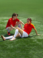 Young soccer player helping teammate stand after fall on field, both wearing red uniforms. Concept...