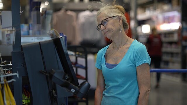 An unhappy, frustrated mature woman stands at the self checkout in a hardware store, looking slightly confused as she processes her purchases with a credit card.
