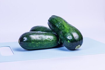Three dark green zucchinis lie on a light blue cutting board. The zucchinis are shiny and fresh, stacked one on top of the other. Cooking, emphasizing the freshness and texture of the zucchinis.