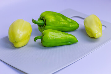 Green and yellow bell peppers are placed on a light gray cutting board. The peppers have a smooth, shiny surface with whole stems. Preparation for cooking, fresh produce on a neutral background.