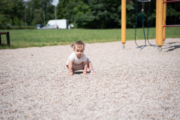young child exploring a sunlit playground, surrounded by pebbles and green trees, kneeling near a...