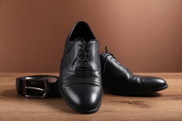 Pair of black leather shoes and belt on wooden surface against brown background, closeup